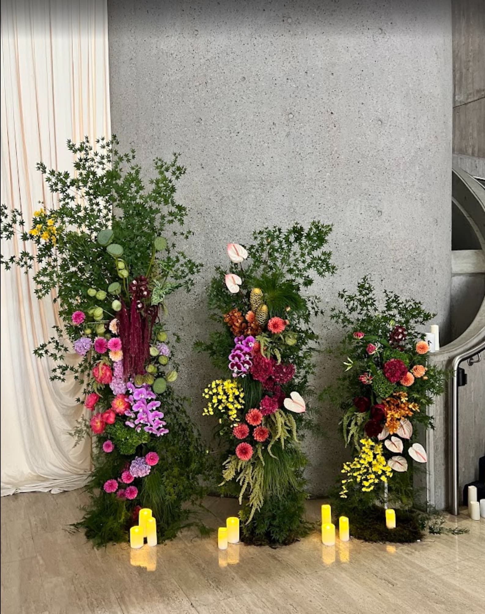 Three tall floral installations against a raw concrete wall, lit by candles