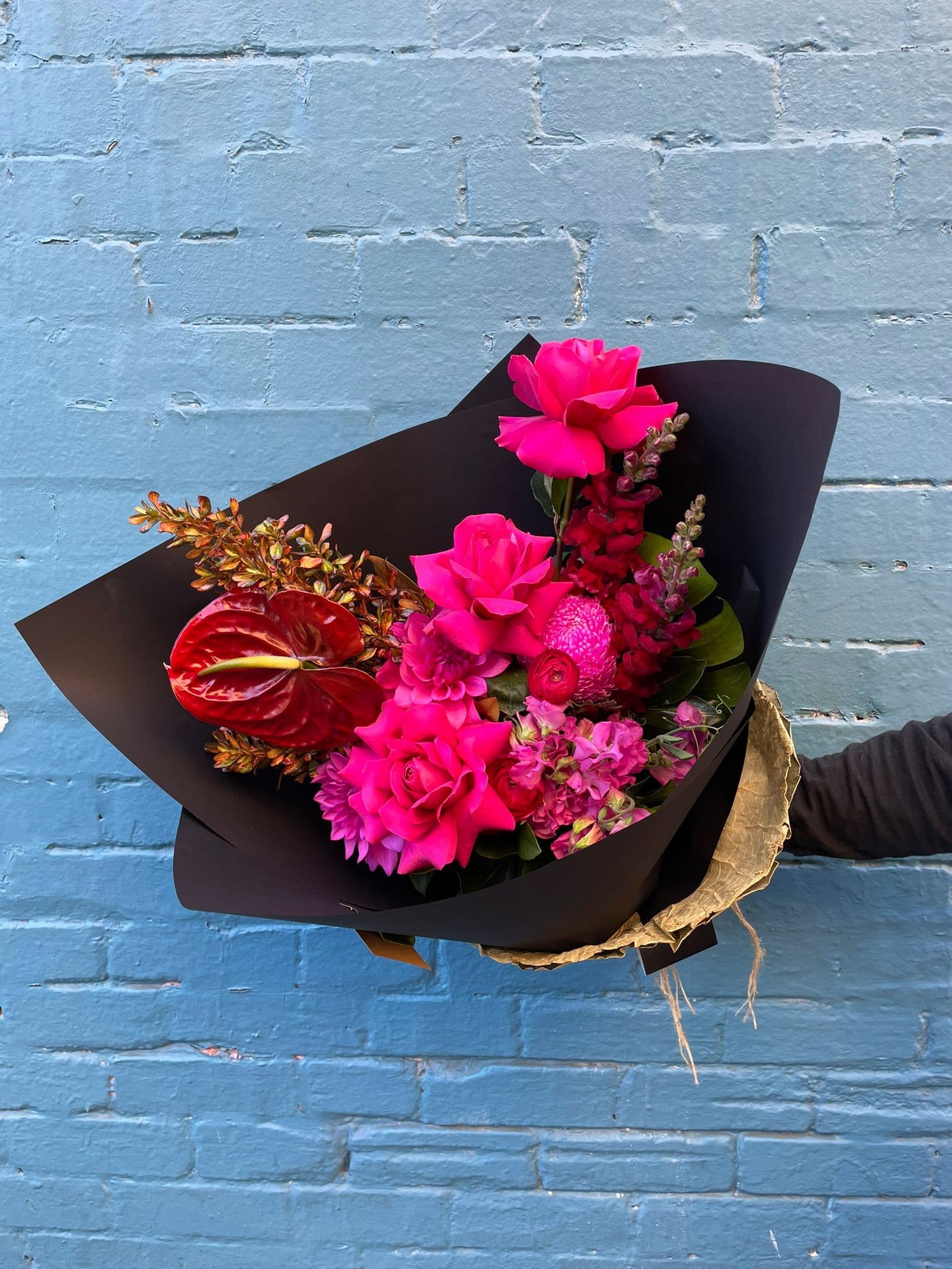 A pink and red hand-tied bouquet in black wrap against a blue wall